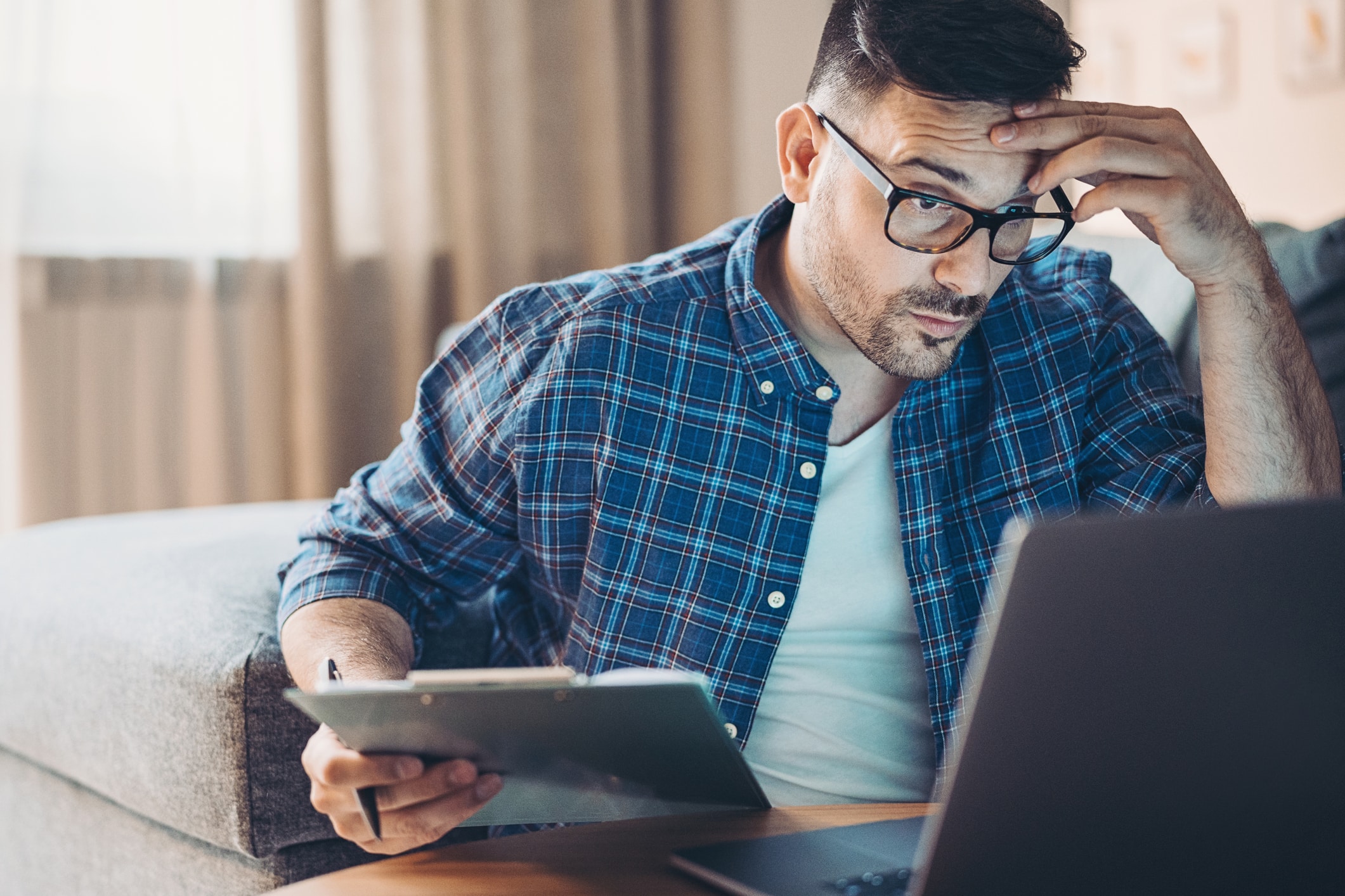 A man with glasses sis working on his laptop with a tablet in his hand, looking stressed or confused.